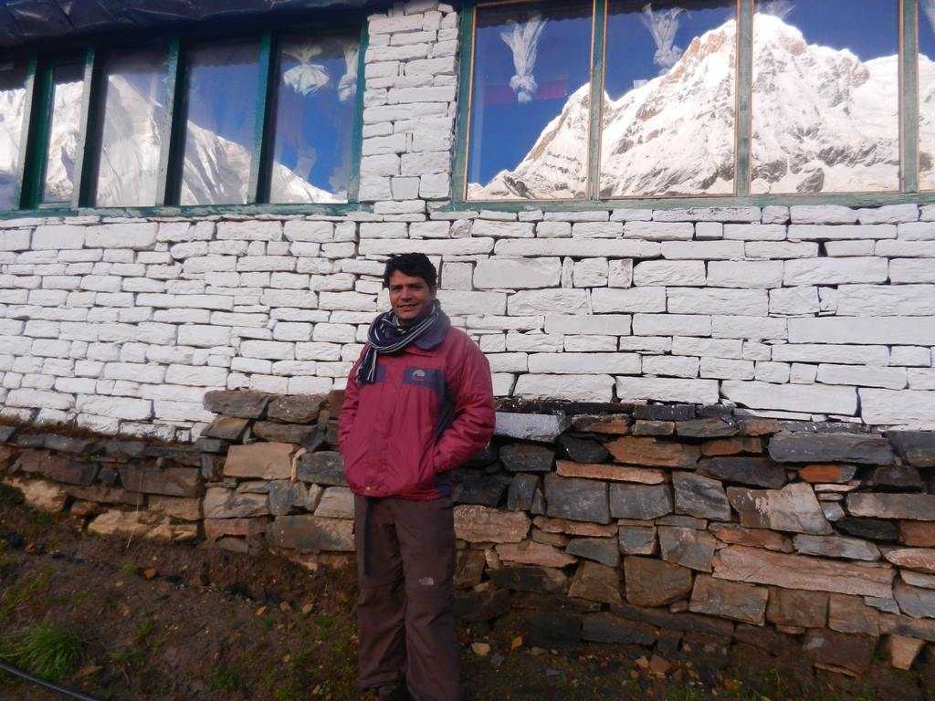 A man infront of a teahouse on whose windows is Annapurna reflecting